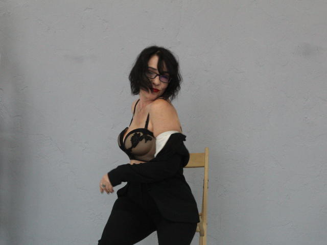 SandraHormas con un sujetador de encaje negro, mostrando senos grandes, pantalones negros y una chaqueta que se desliza del hombro, sentada en una silla de madera, mirando hacia abajo, usando gafas, con cabello negro despeinado..