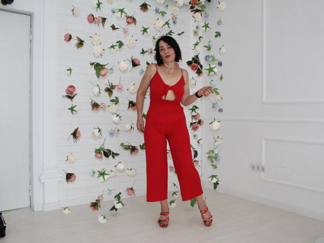 SandraHormas portant une combinaison rouge, découpée et à taille haute, avec des talons rouges, tenant des fleurs, avec des cheveux courts noirs, sur un mur blanc, avec des roses roses, blanches et bleues éparpillées. Les seins sont visibles à travers les découpes..