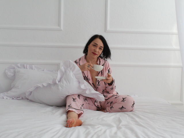 SandraHormas, in pigiama a farfalle rosa, seduta su un letto bianco con cuscino a frange, che tiene una tazza bianca, sorridente, con capelli neri, su uno sfondo di parete bianca, a piedi nudi, carina..