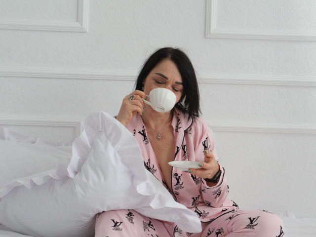 SandraHormas, avec ses cheveux noirs, porte un pyjama rose orné de motifs d'oiseaux noirs, elle boit dans une tasse blanche, tient une soucoupe, est assise sur un lit blanc et regarde vers le bas..