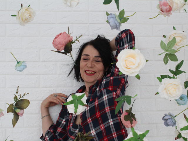 SandraHormas, met zwart haar, lachend, in een rood geruit shirt, met een witte roos in haar hand, omringd door roze, witte en blauwe bloemen..
