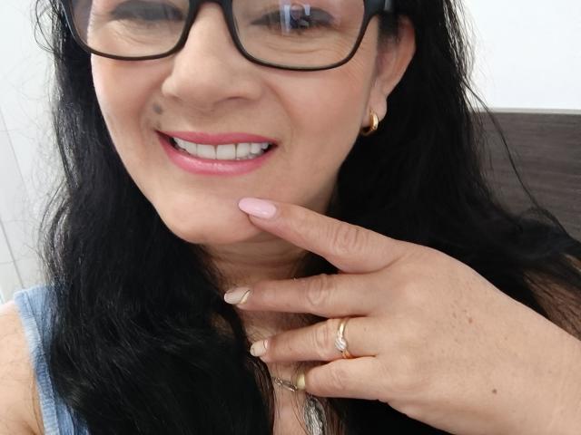 MarcelaExtreme with black hair, glasses, pink lipstick, gold ring, and pink nails, smiling, touching her chin, wearing a blue top, and gold earrings, looking cute.