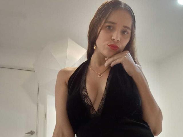 DannaRubyns with red lipstick, black lace halter top showing her tits, gold necklace, and hand near mouth, looking slightly pouty. She has long brown hair and a light skin tone.