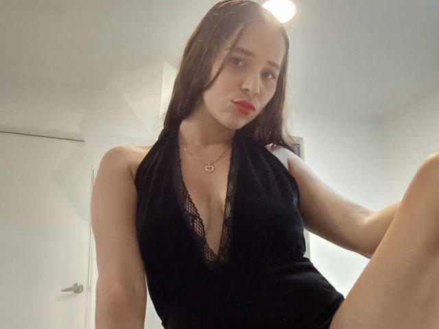 DannaRubyns with long brown hair, red lipstick, black lace halter top, showing deep cleavage, small gold necklace, sitting with one leg up, looking at camera.