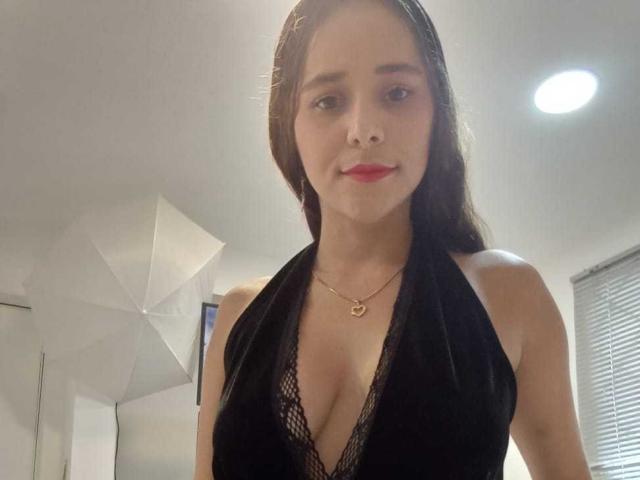 DannaRubyns has medium tits in a black lace halter top, wearing a gold necklace, with straight brown hair and red lipstick, in a softly lit room.