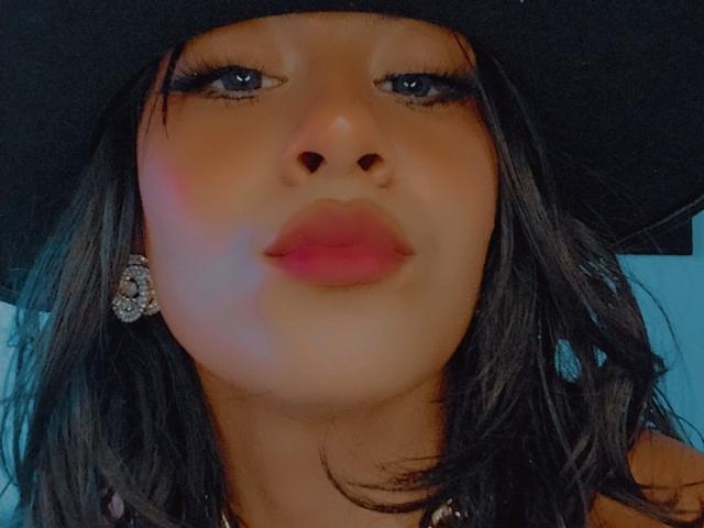 DarlyeCouples Gesicht, mit einem schwarzen Hut, blauen Augen, langen schwarzen Wimpern und rosafarbenen Lippen, hat dunkles, welliges Haar und ein Diamant-Ohrring in ihrem linken Ohr..