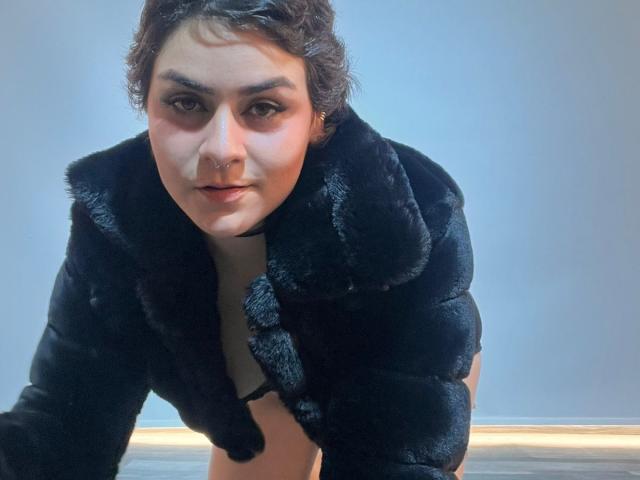 Isabela Cooper, con cabello corto y rizado de color marrón, vestida con un abrigo de piel negro, de rodillas, mirando a la cámara con una ligera sonrisa, mostrando un piercing en la nariz y un maquillaje sutil..