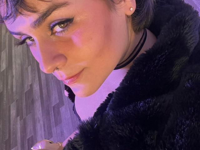 IsabelaCooper has short dark hair, a nose ring, and wears a black furry coat with a black choker, looking up with purple-lit eyes and a slight smile.