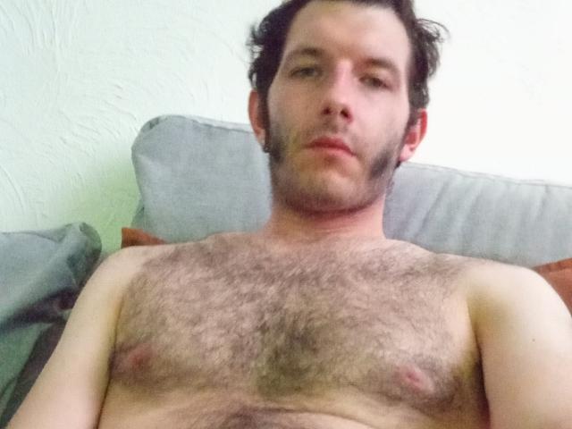 Aure69, sem camisa e com o peito peludo, está deitado em um sofá, mostrando pequenos mamilos levemente eretos e uma expressão facial neutra. Aure69 tem cabelo curto e escuro e uma barba clara..