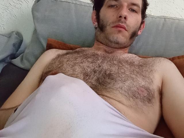 Aure69, senza camicia e con il petto peloso, è sdraiato a letto, coperto da un lenzuolo bianco, e guarda la telecamera con un'espressione leggermente annoiata. Aure69 ha i capelli corti e scuri e dei piercing alle orecchie..