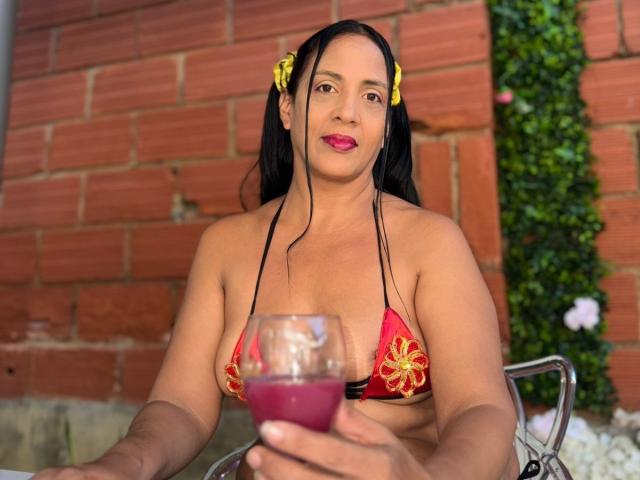 JulietaMilf with medium brown skin, black pigtails, yellow flower clips, red floral bikini, holding purple drink, sitting, smiling, pink lipstick, brick wall background, metal chair.
