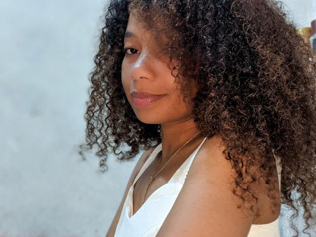MarieBella with curly brown hair, light brown skin, wearing a white tank top, smiling slightly, looking to the side, shiny lips, gold necklace, soft light, upper body.