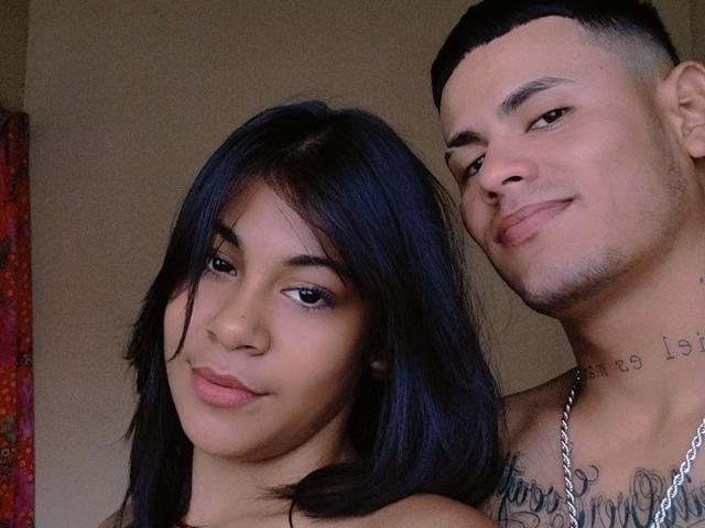 DarlyeCouple mit dunklem Haar, süßem Gesicht und braunen Augen, und DarlyeCouple mit kurzen Haaren, Tattoos und einer Halskette, lächeln gemeinsam. Beide haben hellbraune Haut und sind oben ohne..