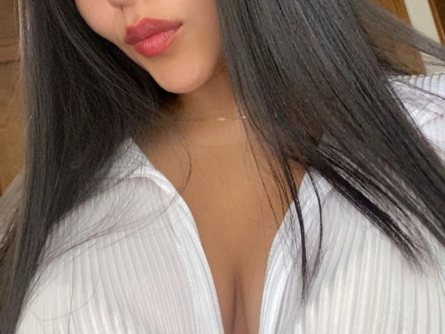 IssaPuasini with long black hair, red lipstick, and a white ribbed shirt showing her big tits and deep cleavage. Her skin is tan. She's wearing a thin necklace.