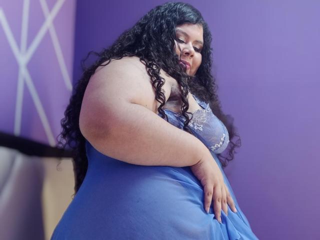 SarahBowie with large, curly black hair, wearing a blue lace nightie, showing her big, fat, pale tits and thick arms, standing against a purple wall.