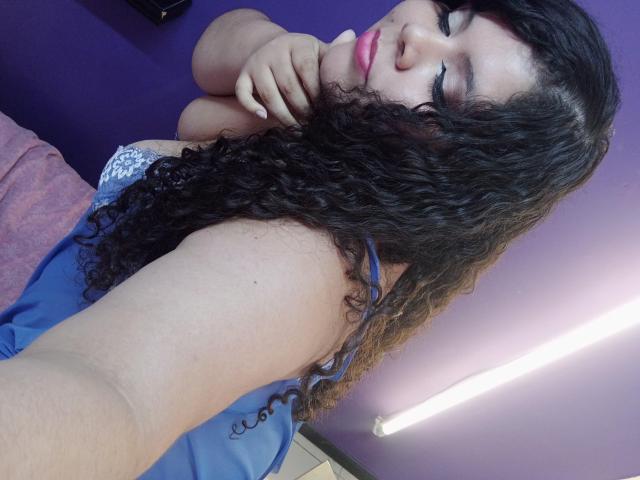 SarahBowie with curly black hair, pink lipstick, blue top, showing her arm and hand, slightly biting her finger, purple wall in background, fluorescent light on right.