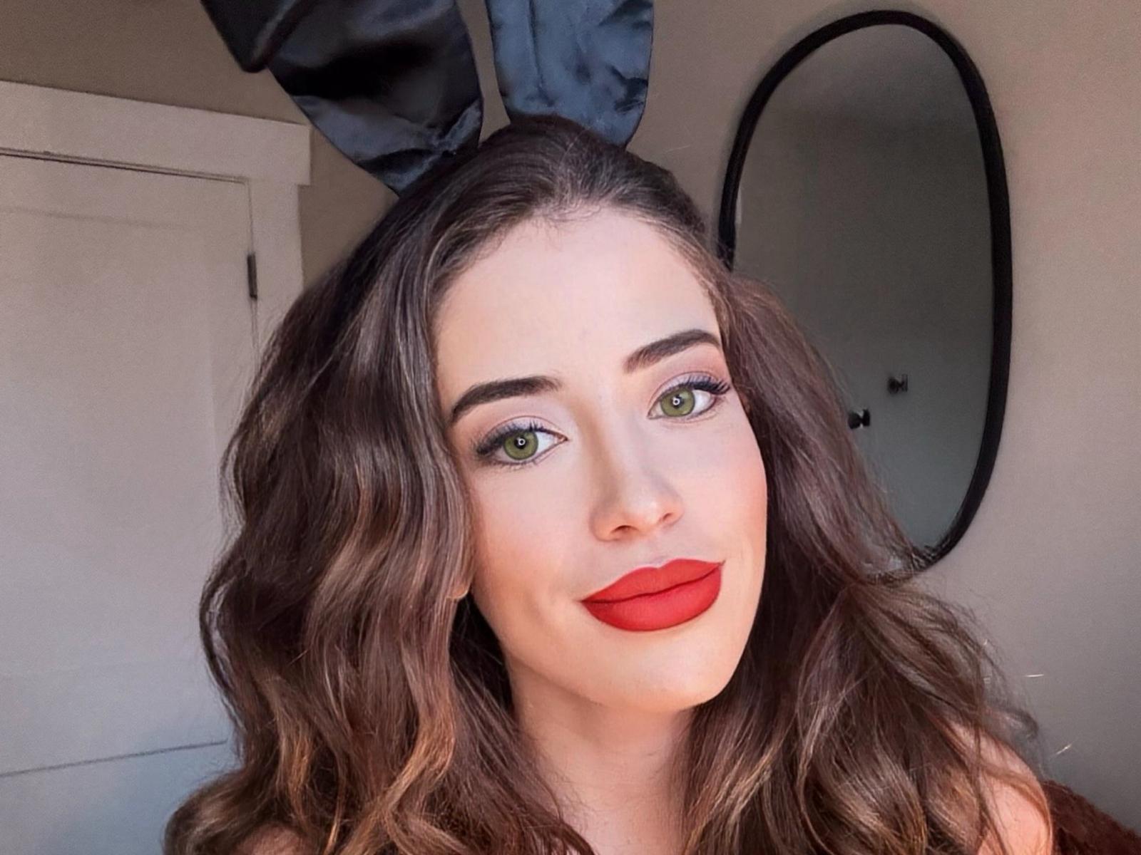 JaseyHott has wavy brown hair, green eyes, and red lipstick. She's smiling with a cute face, and her tits are slightly visible under a brown top.