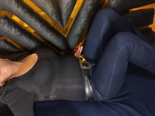 HellenaSexy deitada em um sofá azul escuro, vestindo uma blusa preta justa e jeans azuis, com um cinto com fivela dourada e um cinto preto, com o bumbum ligeiramente levantado, a cabeça parcialmente fora do quadro..