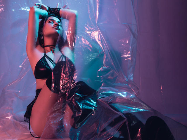 MaiaThunder vestindo lingerie de látex preta brilhante, braços acima da cabeça, sentada em plástico, com iluminação azul e roxa, olhando para cima, luvas pretas, colar e botas que vão até a coxa, pose sexy e confiante..