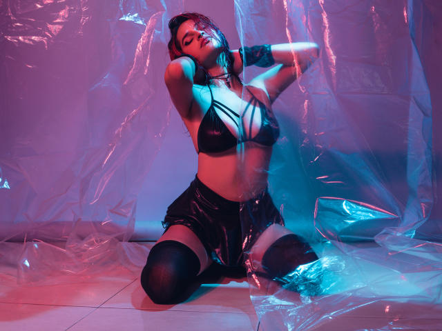 MaiaThunder, agenouillée, porte un soutien-gorge noir à bretelles, un short noir brillant et des bas hauts, ses mains dans ses cheveux bruns, avec des lumières roses et bleues, et du plastique enroulé autour d'elle..