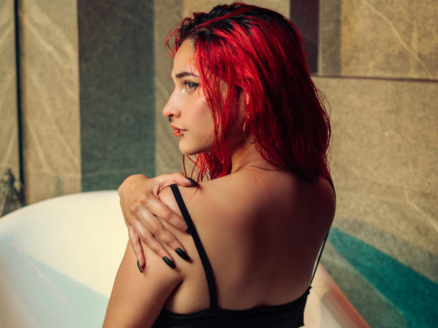 MaiaThunder com cabelo vermelho e molhado, top preto, unhas pretas, braços cruzados, olhando por cima do ombro, em pé em uma banheira branca, mostrando o bumbum e um piercing no nariz..
