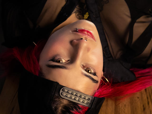O rosto da MaiaThunder virado para baixo, cabelo ruivo, faixa de cabeça preta com tachas de metal, piercings no nariz, batom vermelho, olhos castanhos, blusa preta transparente, piso de madeira, iluminação suave..
