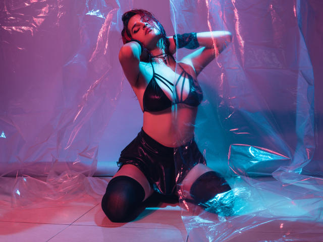 MaiaThunder, con grandi seni, indossa un top di pelle nera con spalline sottili, pantaloncini neri e calze alte fino alla coscia, è inginocchiata su piastrelle, con le mani tra i capelli, circondata da luci al neon blu e rosa..