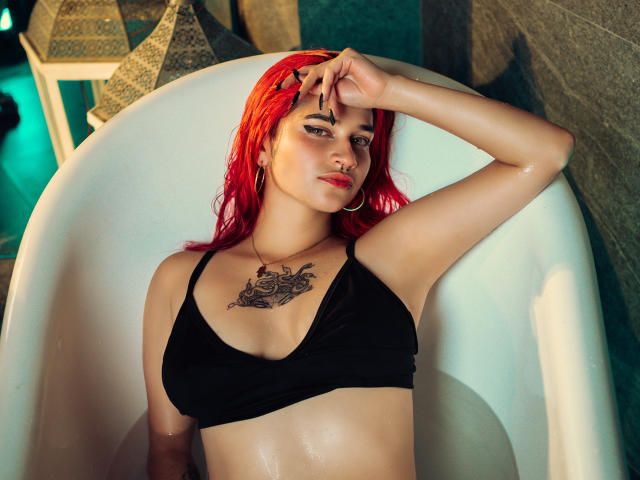 MaiaThunder con i capelli rossi, reggiseno nero, tatuaggio sul petto, mano sulla fronte, seduta in una vasca da bagno bianca, pelle bagnata, piccolo piercing al naso, che guarda la fotocamera, sfondo con piastrelle verdi..