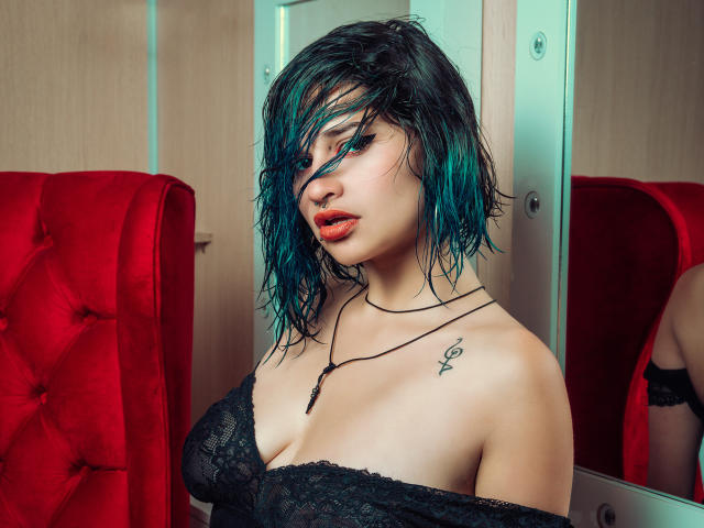 MaiaThunder with teal-dyed, wet-looking hair, wearing black lace off-shoulder top, showing her tits, red lips, nose ring, tattoo on right shoulder, and black necklace. Red chair background.