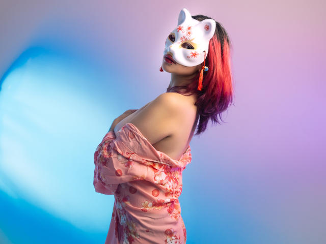 MaiaThunder met een wit vosmasker, haar met een rode en paarse overgang, en een roze kimono met bloemen, haar borsten gedeeltelijk bedekt, haar achterkant nauwelijks zichtbaar, ze ziet er sexy en zelfverzekerd uit..