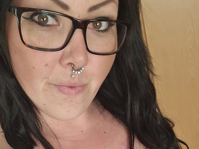 TinyDiamond88-hot heeft zwart, golvend haar, bruine ogen en een neusring met een klein juweeltje. Ze draagt een zwarte bril en heeft lichte sproeten en een lichte glimlach..