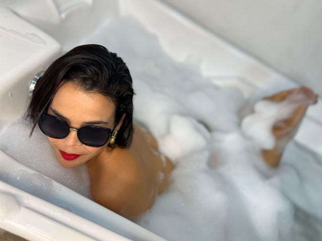 AbriilBeltran with dark hair, red lipstick, and black sunglasses, lying in a bubble-filled bathtub, legs up, exposing her ass, looking at the camera with a playful smile.