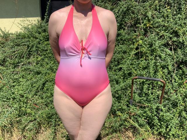 Milf01-hot with pink to pink gradient one-piece swimsuit, hands behind back, standing in front of green bush, visible tan lines, slightly large boobs, small red knot in front.