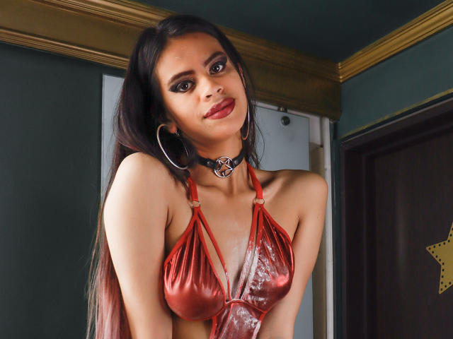 MulanDurand met lang zwart haar, een rode, glanzende leren bustier, een choker met een metalen ring en oorringen, leunt voorover en kijkt naar de camera met rode lippenstift en donkere make-up..