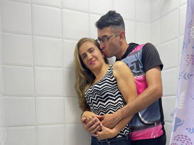 TifanyAndStiven are in a bathroom. Stiven, with glasses and a colorful shirt, kisses Tifany's cheek while holding her hands. Tifany, in a zigzag shirt, smiles.