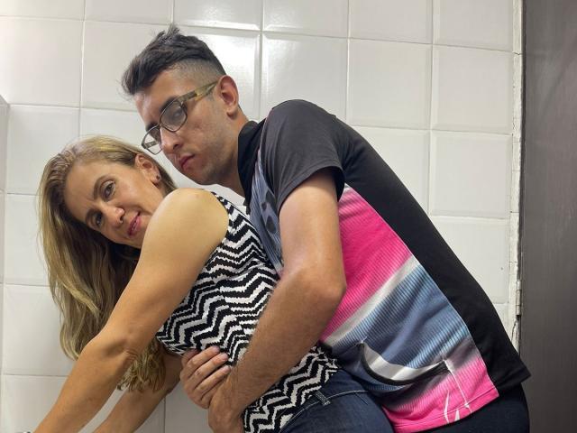 TifanyAndStiven, with long blonde hair and a zigzag top, leans against Stiven, who has dark hair, glasses, and a pink, black, and white shirt, gripping her ass.