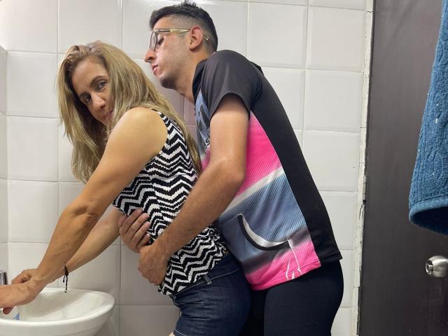 TifanyAndStiven, muscular blonde woman in zigzag top, bent over sink, grabbed by Stiven, wearing glasses, pink and gray shirt, black pants, in white-tiled bathroom.
