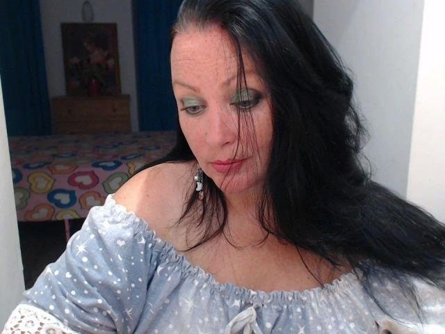ElaineNights-hot with long black hair, blue-green eyeshadow, light skin, and small tits, wearing a blue polka-dot off-shoulder top, looking down with a slight pout in a bedroom.