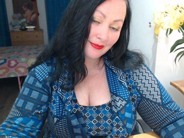 ElaineNights-hot has black wavy hair, yellow eyeshadow, red lipstick, and a blue patterned dress with a low-cut neckline showing her tits. She's sitting with a smile.