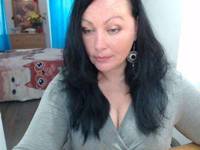 ElaineNights-hot has pale skin, black wavy hair, and is wearing a grey V-neck top showing her tits. She's wearing a sunburst earring, with a slight, sexy smile.