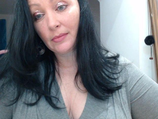 ElaineNights-hot has black hair, pink glitter eyeshadow, gray V-neck shirt, and small tits with a slight cleavage, looking at the camera with a neutral expression.