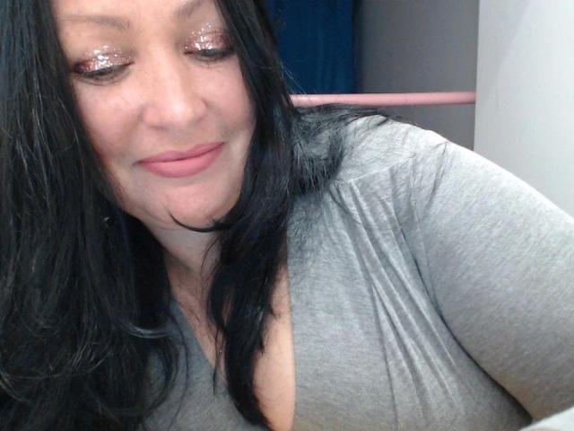 ElaineNights-hot with black hair, wearing a grey top, showing a smile with pink glittery eyes, and visible cleavage, looking down slightly. Her tits are subtly highlighted.