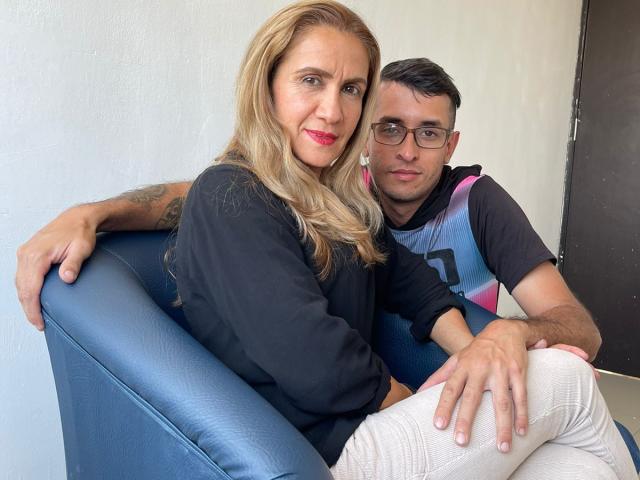 TifanyAndStiven, blonde woman with red lipstick and black shirt, sitting on blue chair, arm around tattooed man in blue and pink shirt, white pants, both smiling.