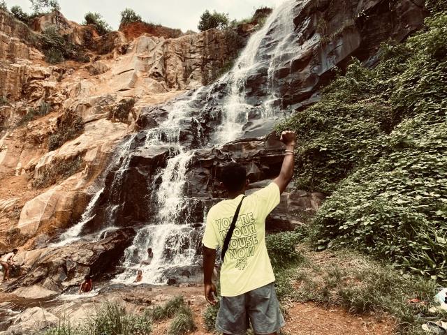 Dzeus, qui a la peau foncée, portant une chemise jaune et un short bleu, se tient debout, bras levé, face à une cascade rocheuse. De petites personnes se trouvent près de l'eau..