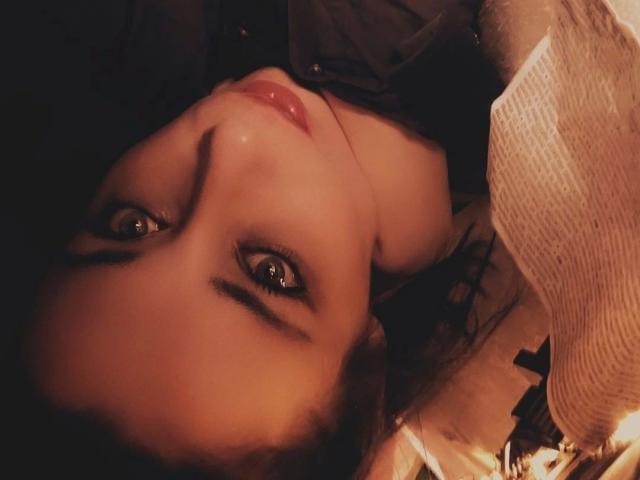 Mary-Licious has brown eyes, shiny red lips, and a slightly upturned nose, lying down with dark hair and a black garment. Soft, warm lighting highlights her face.