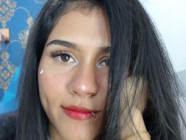 AnnieDarling with dark hair, brown eyes, and a small nose ring, smiling slightly with red lipstick, wearing a gold bracelet, and a tiny white mark on her right cheek.