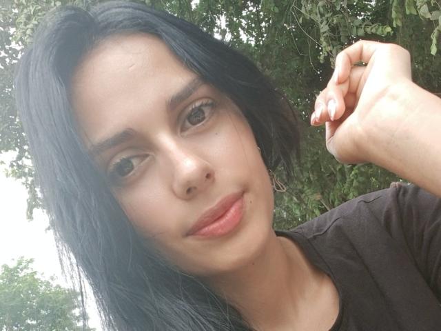 AnaisHilton has black hair, brown eyes, and a small nose boof, wearing a black shirt, with a slightly parted lip and a hand touching her head.