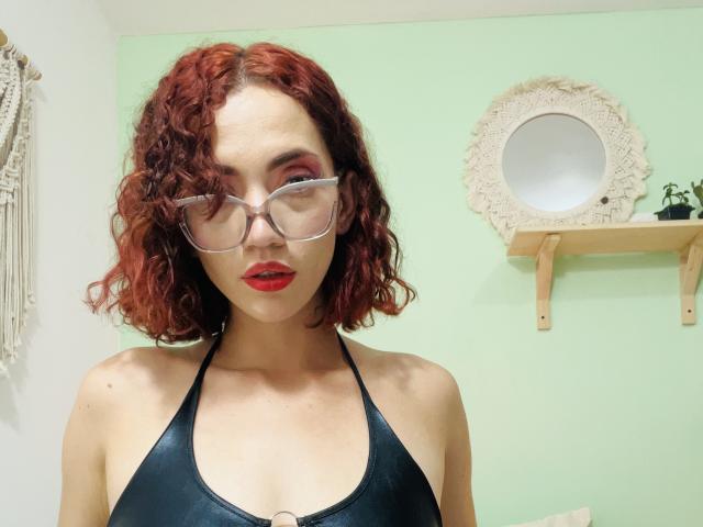 AmandaGougis with curly red hair, wearing clear glasses and a black halter top, showing her tits, red lipstick, and a confident expression.