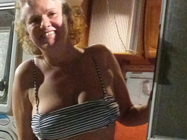Jutta1-hot with curly blonde hair, smiling, wearing a striped bra that barely covers her tits, standing in a dimly lit room, looking into a mirror.
