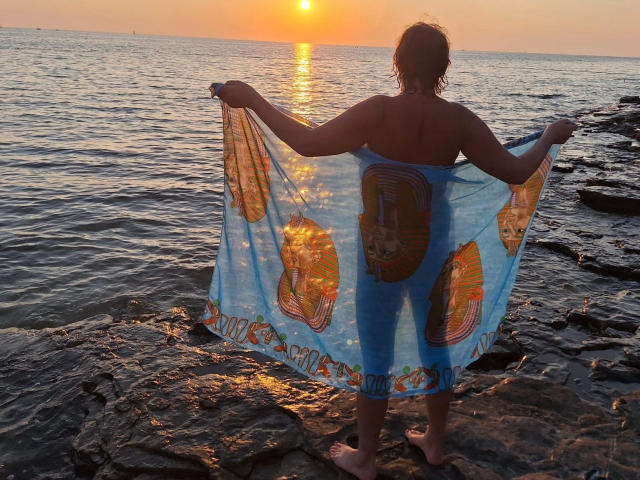 Jutta1-hot stands on rocky shore, back to camera, holding up a blue, patterned, see-through sarong, sunlight reflecting on water, her ass slightly visible, wavy brown hair.